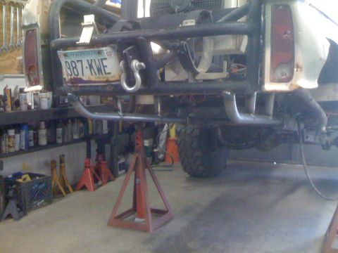 1966 Ford Bronco - The rear of a custom off-road vehicle is supported by jack stands in a garage, with tools and shelves visible in the background.