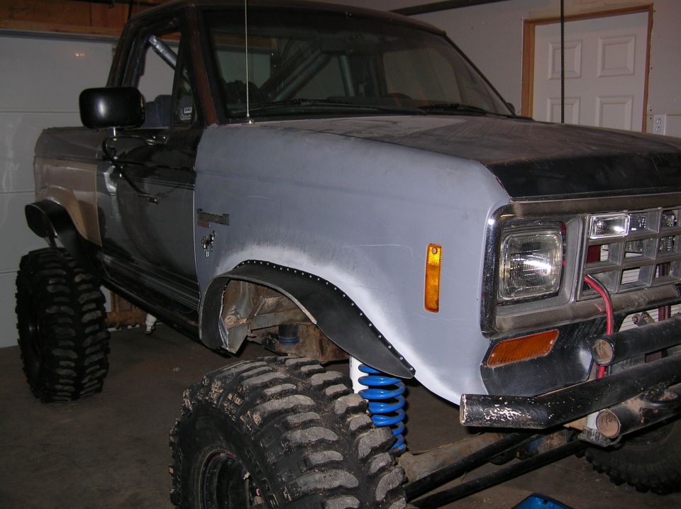 Lifted 1980s Ford 4x4 with primer-gray fender, blue coils and mud tires in a garage