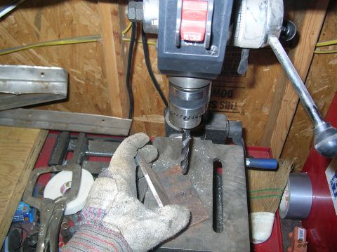 1985 Ford Bronco II - A drill press is used to drill a hole in a metal plate, with a gloved hand holding the workpiece.