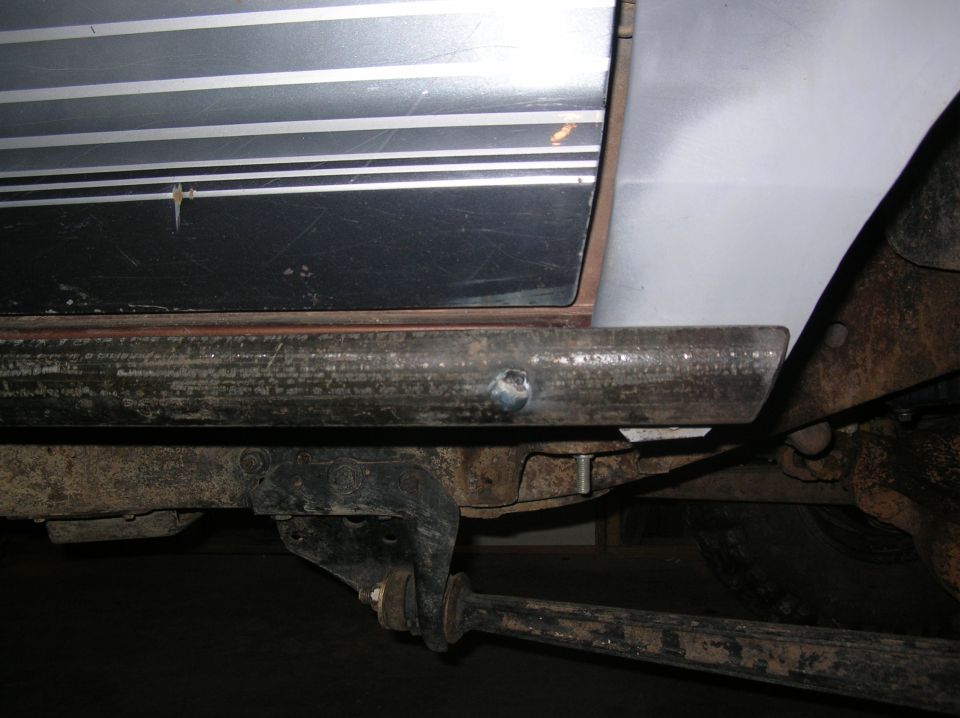 Bare steel rock slider bolted to rocker panel of silver truck, frame and links below