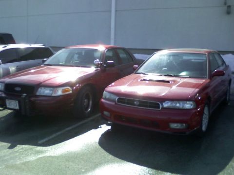 1997 Subaru Legacy - A red 1997 Subaru Legacy sedan is parked next to another vehicle, showing its front grille and headlights.