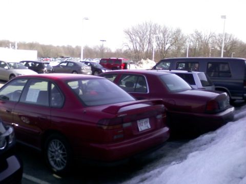 1997 Subaru Legacy - A red 1997 Subaru Legacy sedan is parked in a lot with other vehicles, showing its rear profile and spoiler.