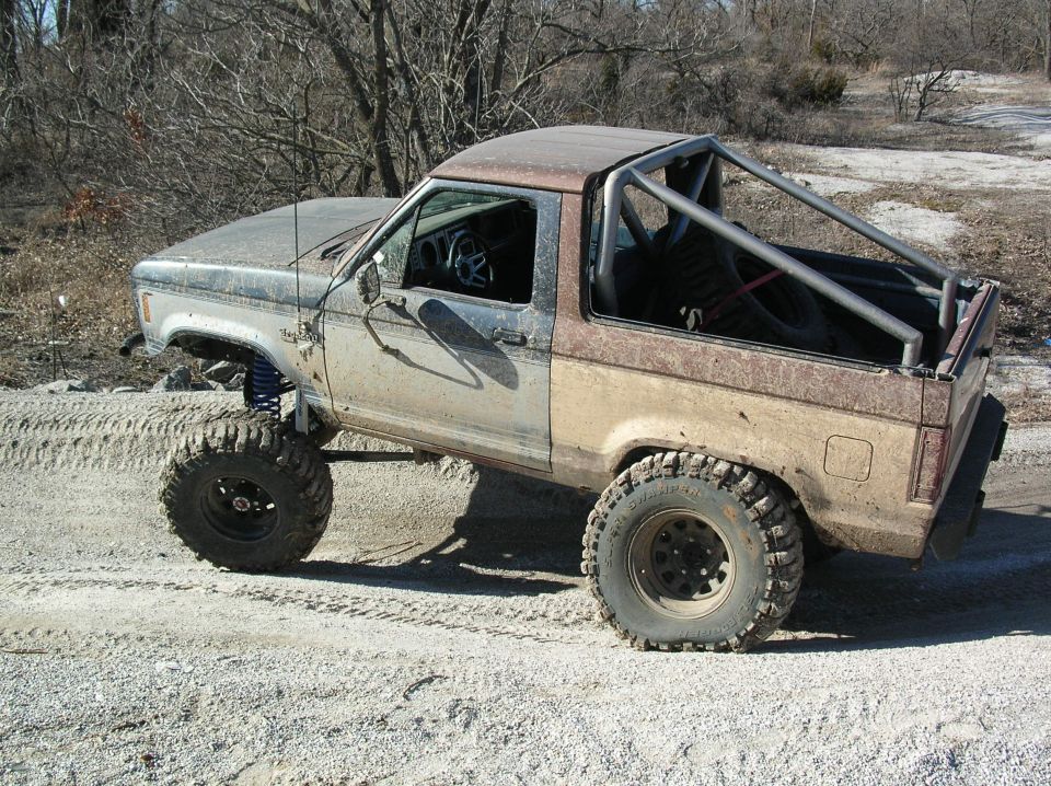 Lifted, muddy blue Ford Bronco II on large off-road tires with roll cage and no top, side view