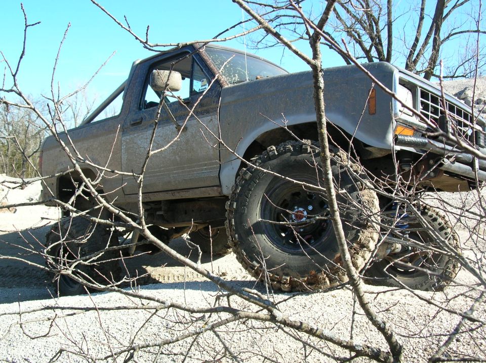 Muddy tan lifted Ford truck on large off-road tires seen behind bare branches