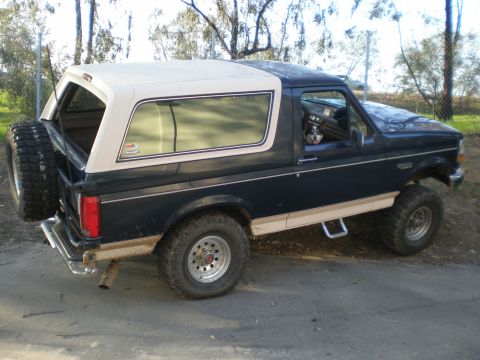 1993 Ford Bronco - A 1993 Ford Bronco is seen from the rear quarter angle, a dark blue body and a tan removable top with a spare tire mounted externally.
