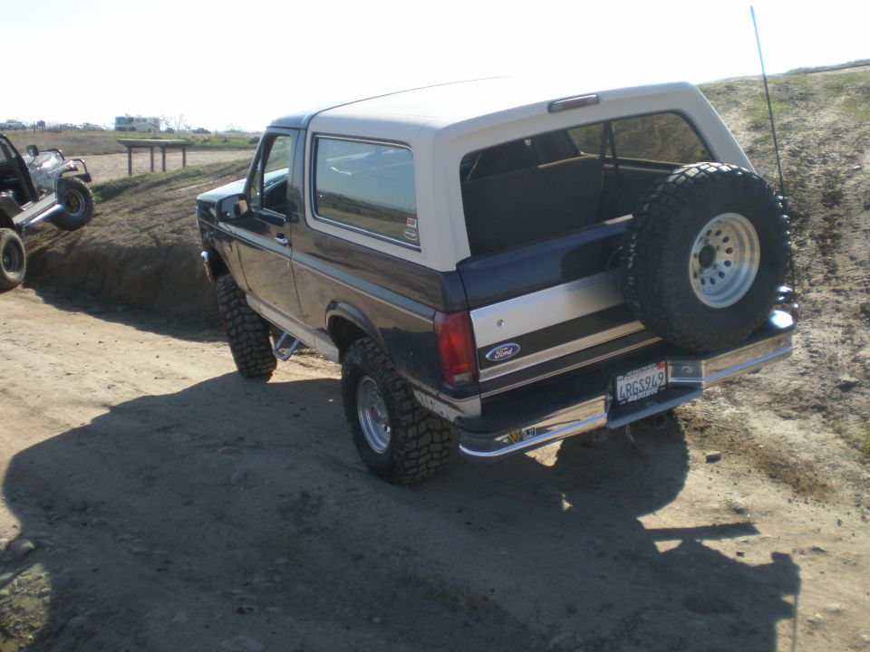 Rear view of a 1993 Ford Bronco with a spare tire mounted on the back, driving on a dirt road. Another ATV is visible on a hill