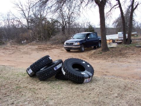 1990 Ford C-150 (Centurion Conversions) - A dark blue pickup truck is parked outdoors near a pile of four large off-road tires.