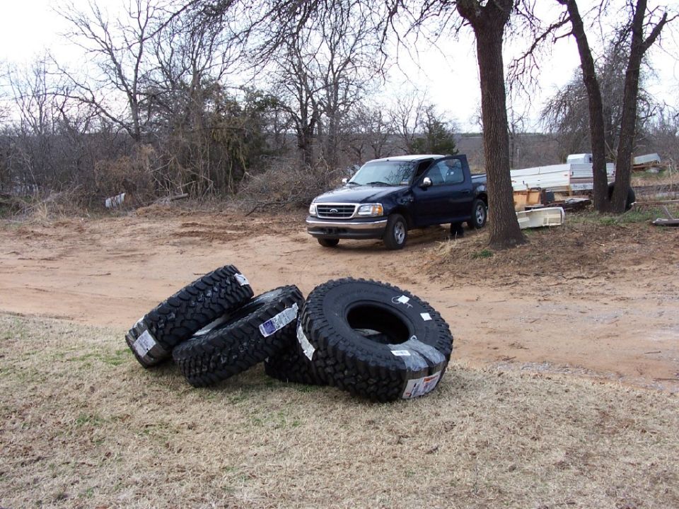 Dark blue pickup truck, 1999 model year, with a tan lower body trim. Four large off-road tires are stacked in the foreground