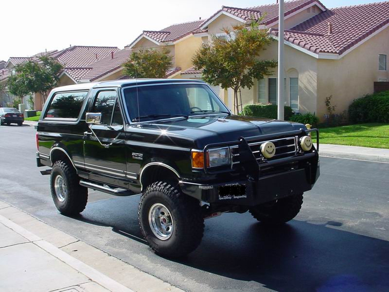 Black 1987 Ford Bronco with custom grille guard and smiley face lights, parked on a suburban street
