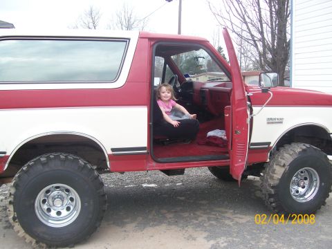 1988 Ford Bronco - Child sits in the driver's seat of a red and white 1988 Ford Bronco with the passenger door open.