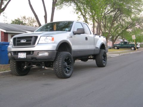 1979 Ford Bronco - A lifted silver Ford F-150 pickup truck is parked on the street, with another green pickup truck in the background.