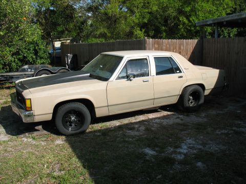 1985 Ford LTD Crown Victoria - A tan sedan with a contrasting black hood sits parked in a grassy area next to a wooden fence.