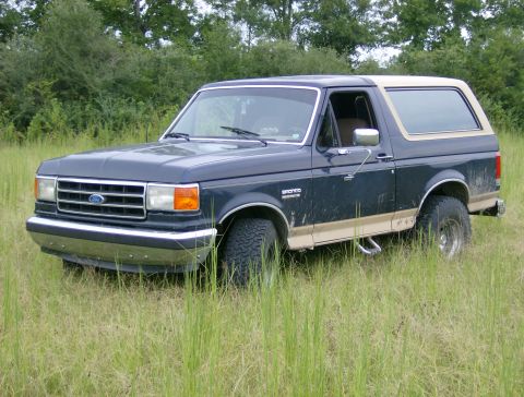 1990 Ford Bronco - A dark blue Ford Bronco with tan trim sits in a grassy field with trees in the background.