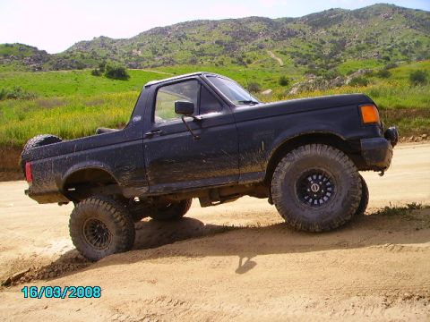 1990 Ford Bronco - This lifted Ford Bronco is navigating a dusty, off-road trail with green hills in the distance.