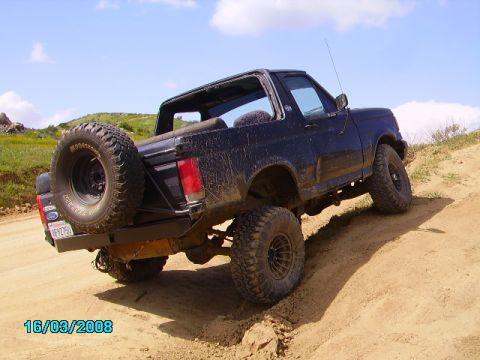 1990 Ford Bronco - A black 1990 Ford Bronco is shown from the rear-side angle, driving on a dirt path with a spare tire mounted externally.