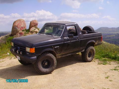 1990 Ford Bronco - 1990 Ford Bronco parked on a dirt hill with large rocks and a distant town.