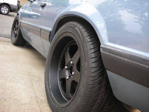 1990 Ford Mustang - A close-up shot of a car's side profile, focusing on its wheel and fender area, with another vehicle visible in the background.
