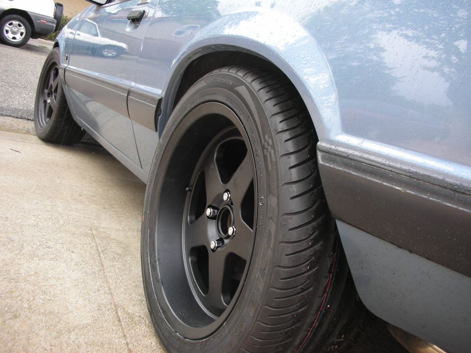 Car side view showing black five-spoke rims and tires. Another car is parked behind it