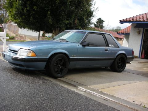 1990 Ford Mustang - A blue coupe with black wheels is parked on a wet driveway.