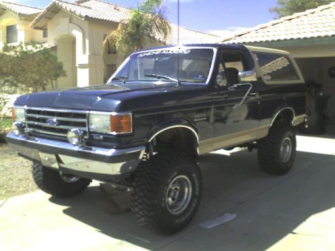 1991 Ford Bronco - This lifted Ford Bronco features a dark blue body with tan accents and prominent off-road tires.