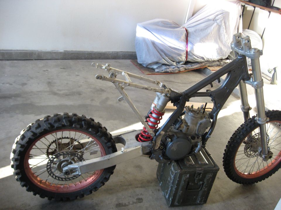 Disassembled dirt bike frame with engine and red shock, resting on an ammo can in a garage.