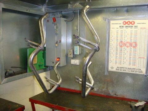 1996 Ford Bronco - A pair of fabricated tubular exhaust headers hanging by wire in a shop, freshly welded and in bare metal, in front of an electrical panel a…