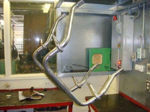 1996 Ford Bronco - Bare bent steel tube brackets with welded tabs hang on wires in a shop, raw metal still unpainted before finishing. Behind them are a green…