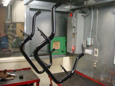 1996 Ford Bronco - Four freshly powder-coated gloss black bent tubular brackets with welded mounting tabs hang on wire hooks in an industrial shop next to an…