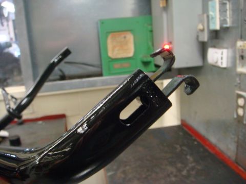 1996 Ford Bronco - Close-up of a freshly coated glossy black steel bracket with a slotted mounting hole and hooked arms, hanging on a rack in an industrial po…