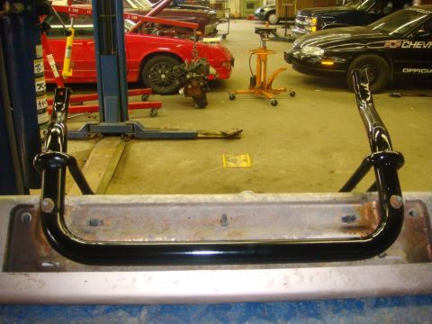 1996 Ford Bronco - Close-up of a gloss-black tubular push bar or grille guard bolted to a bare, surface-rusted bumper support inside a working shop bay. Behin…