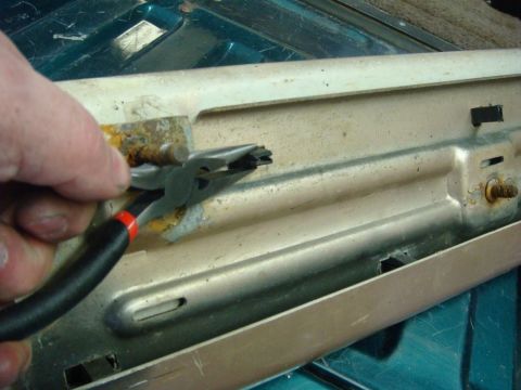 1996 Ford Bronco - A hand uses needle-nose pliers to work a corroded pin on a beige-painted stamped-steel panel with slotted mounting holes, rust blooming aro…