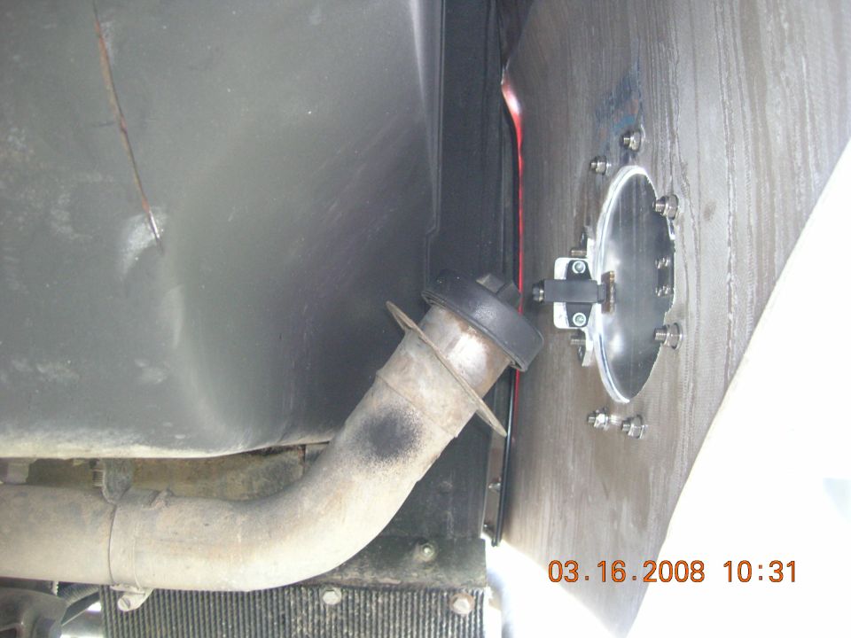Close-up of rusty fuel filler neck through body panel with bolted oval access plate on tank