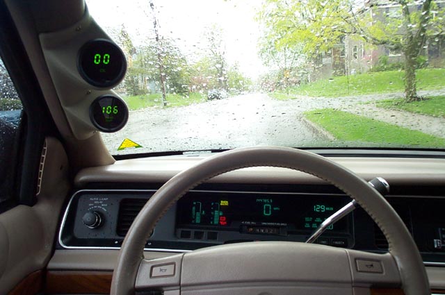 Driver's view of tan interior with digital dash and A-pillar gauge pod, rainy street through windshield