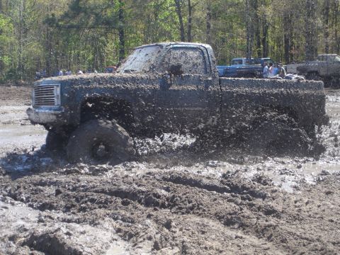 1989 Ford Bronco - This heavily mud-caked pickup truck is stuck deep in a muddy mire, with other vehicles visible in the background.