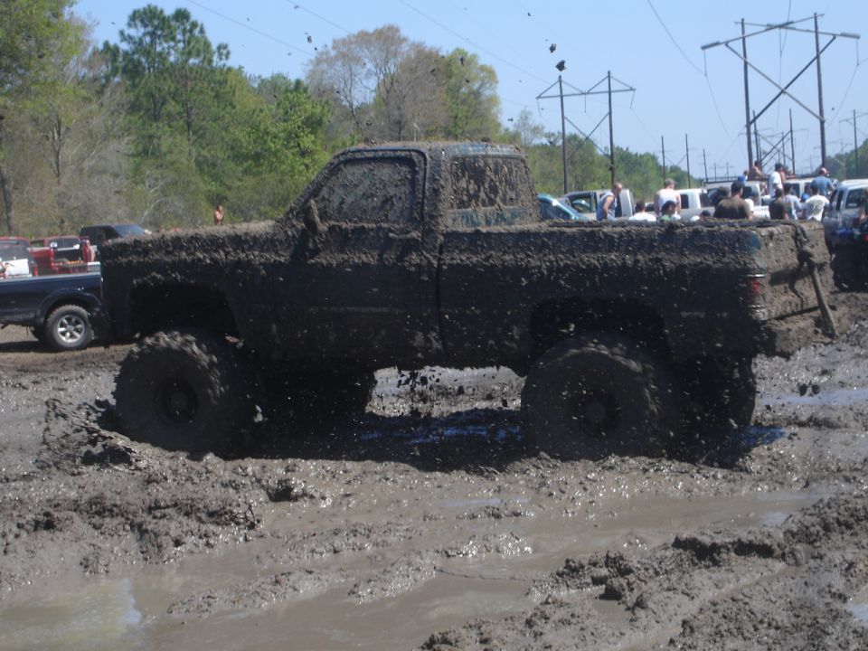Mud-covered pickup truck driving through a mud pit with other vehicles and spectators in the background