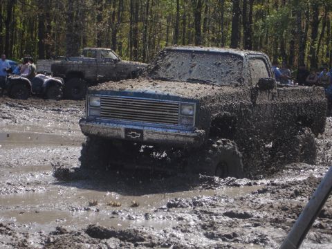 1989 Ford Bronco - A pickup truck navigates a muddy obstacle course, with another truck and ATVs visible in the background.