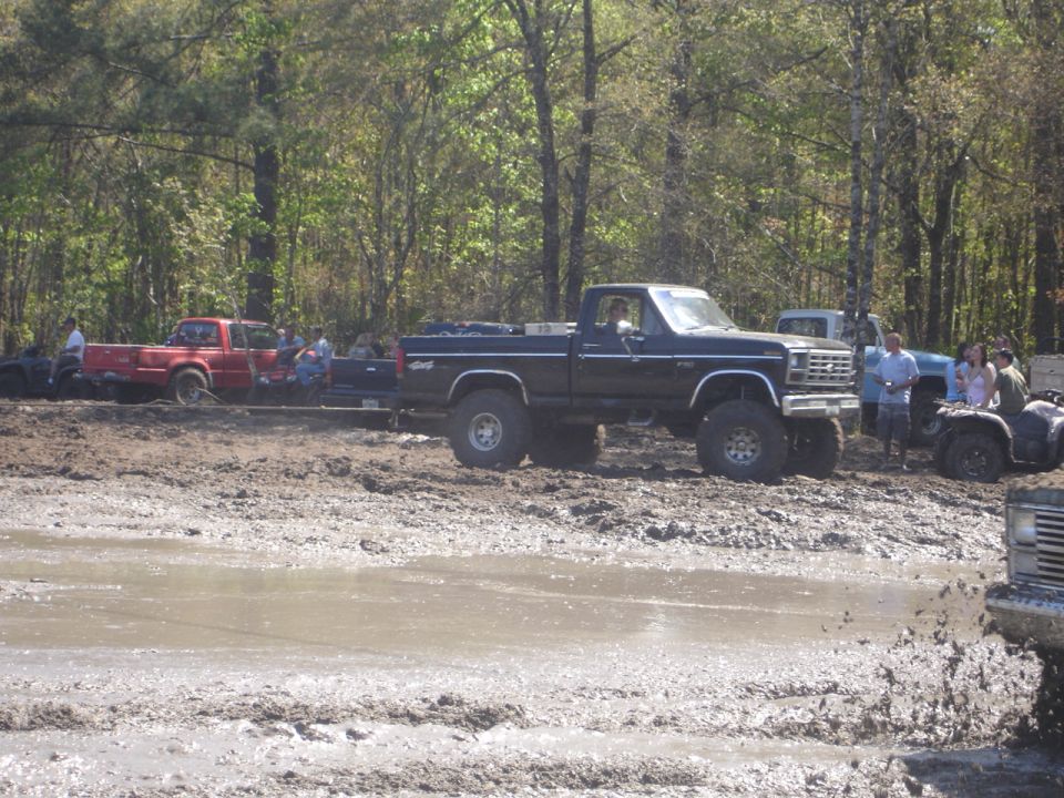 Lifted black pickup truck with 4x4 badging in mud, other trucks and ATVs visible in background
