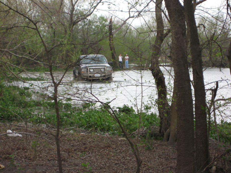 1996 Ford Bronco driving through shallow water with two people standing nearby