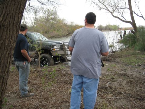 1996 Ford Bronco - A black Ford Bronco is stuck in muddy water with two men observing.