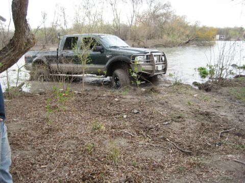 1996 Ford Bronco - Green crew cab pickup truck stuck in mud near a body of water.