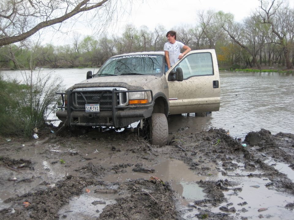 Mud-covered tan Ford F-250 Super Duty with open driver door stuck in mud near water