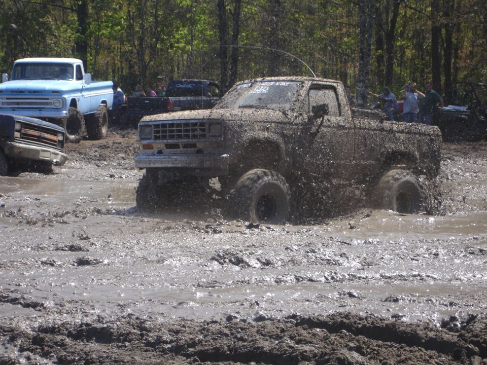 Mud-covered pickup truck with lifted suspension driving through a mud pit. Other trucks visible in background