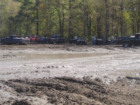 1989 Ford Bronco - Multiple trucks and ATVs are gathered in a muddy off-road area, surrounded by trees.