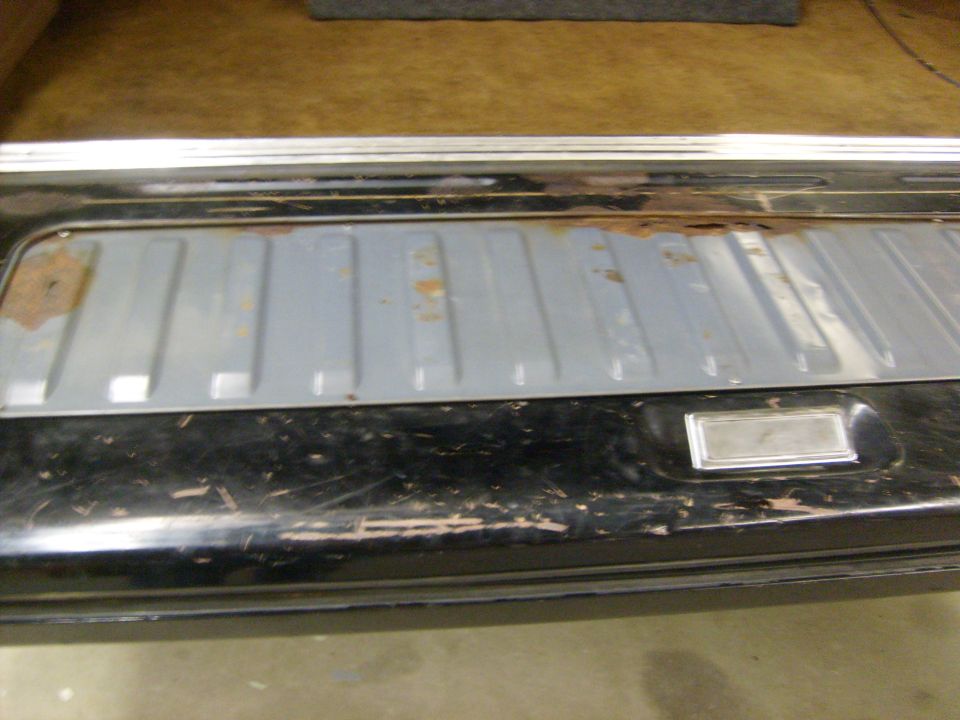 Close-up of a vehicle's rear cargo area, showing a gray ribbed panel with rust spots and a black painted surface with scratches. There's a chrome trim piece on the right