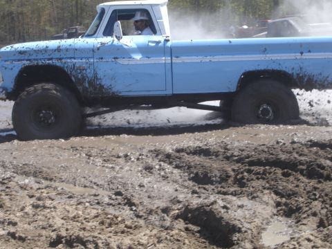 1989 Ford Bronco - A light blue pickup truck with oversized tires is stuck deep in mud, with another vehicle visible in the background.