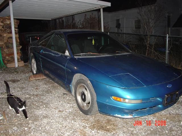 Faded blue Ford Probe on gravel under a carport at dusk with a cat walking past
