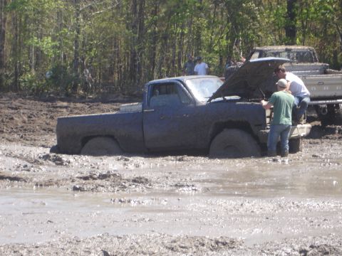 1989 Ford Bronco - Muddy pickup truck with hood up, stuck in a mire with another vehicle in the background.