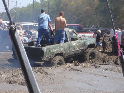 1989 Ford Bronco - A muddy scene with a green pickup truck deeply stuck in mud, while several people stand in its bed and around it.
