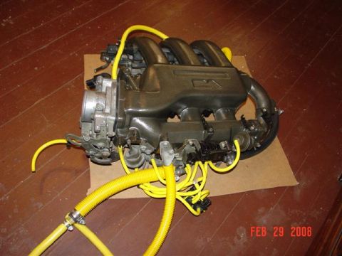 1993 Ford Probe - A close-up of an engine intake manifold with yellow hoses attached, resting on a cardboard piece on a wooden floor.