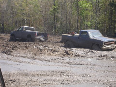 1989 Ford Bronco - Two pickup trucks are stuck in deep mud with trees in the background.